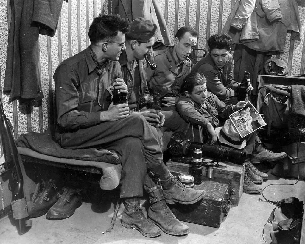 Five U.S. Army soldiers in combat fatigues sitting together during World War II, with photography equipment visible on the ground.